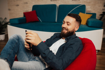 Young attractive smiling guy is browsing at his phone funny video and smiling, sitting at home on the cozy armchair. 