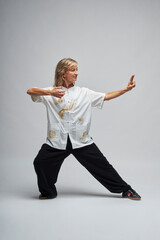 Mature blonde woman practicing Chi kung and Tai Chi on a white background. She wears a traditional white chinese Tai Chi jacket, black trousers and black shoes with ying yang symbol