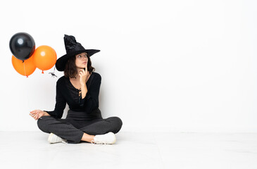Young witch holding black and orange air balloons sitting on the floor having doubts while looking up