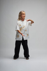 Mature blonde woman practicing Chi kung and Tai Chi on a white background. She wears a traditional white chinese Tai Chi jacket, black trousers and black shoes with ying yang symbol