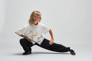 Mature blonde woman practicing Chi kung and Tai Chi on a white background. She wears a traditional white chinese Tai Chi jacket, black trousers and black shoes with ying yang symbol