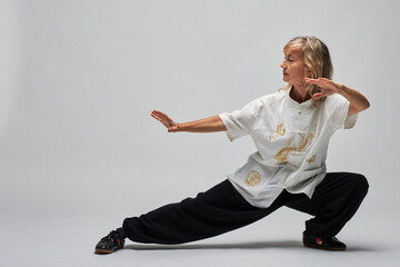 Mature blonde woman practicing Chi kung and Tai Chi on a white background. She wears a traditional white chinese Tai Chi jacket, black trousers and black shoes with ying yang symbol