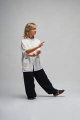Mature blonde woman practicing Chi kung and Tai Chi on a white background. She wears a traditional white chinese Tai Chi jacket, black trousers and black shoes with ying yang symbol