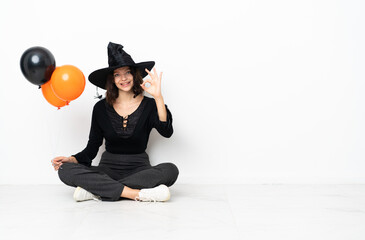 Young witch holding black and orange air balloons sitting on the floor showing ok sign with fingers