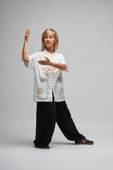 Mature blonde woman practicing Chi kung and Tai Chi on a white background. She wears a traditional white chinese Tai Chi jacket, black trousers and black shoes with ying yang symbol