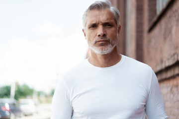 Portrait of a Grey-haired Mature handsome man in jeans and white t-shirt leaning to the old wall.