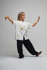 Mature blonde woman practicing Chi kung and Tai Chi on a white background. She wears a traditional white chinese Tai Chi jacket, black trousers and black shoes with ying yang symbol