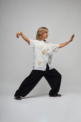 Mature blonde woman practicing Chi kung and Tai Chi on a white background. She wears a traditional white chinese Tai Chi jacket, black trousers and black shoes with ying yang symbol