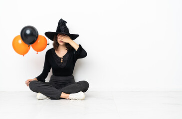 Young witch holding black and orange air balloons sitting on the floor covering eyes by hands. Do not want to see something