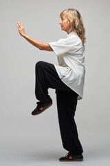 Mature blonde woman practicing Chi kung and Tai Chi on a white background. She wears a traditional white chinese Tai Chi jacket, black trousers and black shoes with ying yang symbol