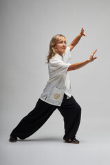 Mature blonde woman practicing Chi kung and Tai Chi on a white background. She wears a traditional white chinese Tai Chi jacket, black trousers and black shoes with ying yang symbol