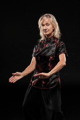 Mature blonde woman practicing Tai Chin Chen style in a black background wearing a traticional chinese black jacket with red decoration, black trousers and traditional shoes with ying yang symbol