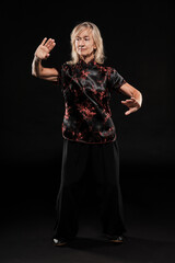 Mature blonde woman practicing Tai Chin Chen style in a black background wearing a traticional chinese black jacket with red decoration, black trousers and traditional shoes with ying yang symbol