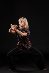 Mature blonde woman practicing Tai Chin Chen style in a black background wearing a traticional chinese black jacket with red decoration, black trousers and traditional shoes with ying yang symbol
