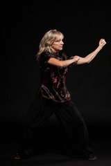 Mature blonde woman practicing Tai Chin Chen style in a black background wearing a traticional chinese black jacket with red decoration, black trousers and traditional shoes with ying yang symbol