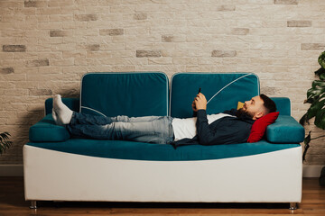 Full length relaxed young bearded  man lying on comfortable blue couch, enjoying spending weekend leisure time with phone, watching funny videos.