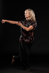 Mature blonde woman practicing Tai Chin Chen style in a black background wearing a traticional chinese black jacket with red decoration, black trousers and traditional shoes with ying yang symbol