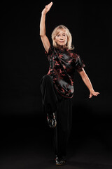 Mature blonde woman practicing Tai Chin Chen style in a black background wearing a traticional chinese black jacket with red decoration, black trousers and traditional shoes with ying yang symbol