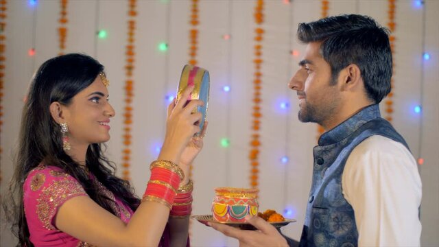 Newly-wed Indian couple celebrating Karwa Chauth festival in traditional wear. Beautiful wife happily looking at the moon and then to her handsome husband through a sieve - Karwa Chauth rituals