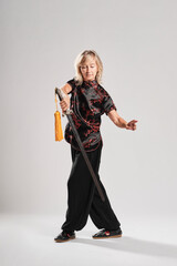 Fototapeta premium Mature blonde woman practicing Tai Chin with sword in a white background wearing a traditional chinese black jacket with red chinese decoration, black trousers and chinese shoes with ying yang symbol