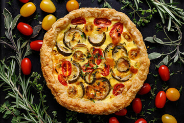 Vegetarian puff pastry quiche with zucchini, tomatoes and herbs on a black background top view