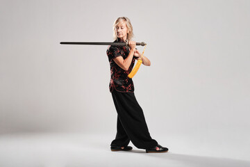 Mature blonde woman practicing Tai Chin with sword in a white background wearing a traditional chinese black jacket with red chinese decoration, black trousers and chinese shoes with ying yang symbol