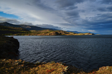 Red stone rocks bay of lake baikal with yellow dry grass in autumn. Blue water with islands, clouds Sunset light. Shadows