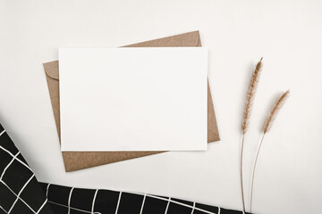 Blank white paper on brown paper envelope with Bristly foxtail dry flower and and Black cloth with White grid pattern. Mock-up of horizontal blank greeting card. Top view of Craft envelope on white 