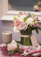 Florist equipment with flowers on white-pink table, top view. Bouquet, candle, secateur, business card of florist shop.