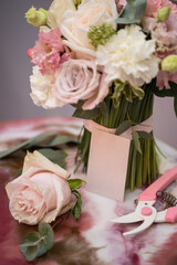 Florist equipment with flowers on white-pink table, top view. Bouquet, candle, secateur, business card of florist shop.