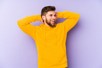 Young man isolated on purple background feeling confident, with hands behind the head.