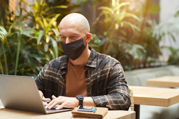 Mature man in protective mask sitting at the table and working on laptop in cafe