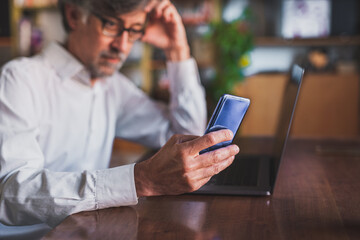 thoughtful businessman wearing glasses and sitting at his laptop, working from home in quarantine, using smartphone to connect and send message, focus on hand and smartphone, blurred background