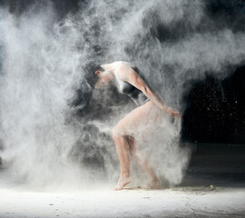 beautiful caucasian woman in a black bodysuit with a sports figure is dancing in a white cloud of flour