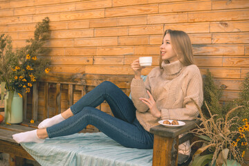 Autumn mood. A smiling girl in a cozy brown sweater and blue jeans sits on a bench and drinks coffee with croissants
