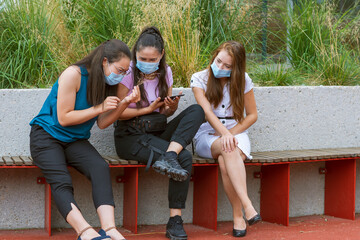 young people wearing a protective mask, sitting and watching on a phone on the playground. Conceptual Coronavirus quarantine and multiracial people having fun together.