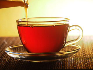 A glass Cup of tea is filled with honey from a glass container.