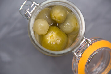 Pickled green small tomatoes in a glass jar with a lid on a gray background. Salted cherries, home-made, olives in brine.