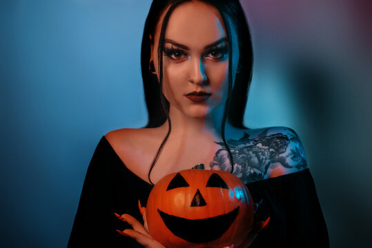 Scary Halloween witch looks into the camera and holds a pumpkin. Fortune teller in neon. Brunette women with tattoo and piercings looking at the camera.
