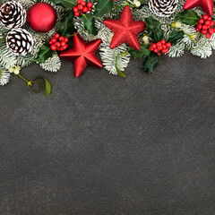 Festive Christmas background border with red star bauble decorations, snow covered fir, holly & winter flora. Composition for the Xmas & New Year holiday season.
