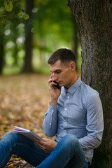 Portrait of a young man in an autumn park, near a tree, outdoors on a natural green background. Serious businessman talking on the phone. Business concept. Selective focus
