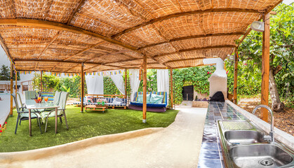 Exterior panorama of a backyard with a lounge area in a relaxing holiday villa with a sofas, pillows,table, chairs,barbecue, grass, garden and a pool