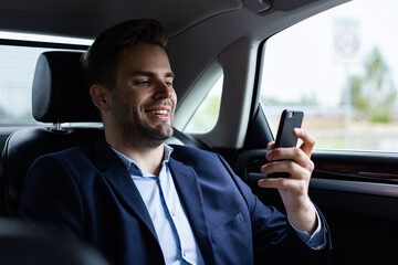 Close up portrait of young man in suit holding smartphone while sitting on the back seat of the car. Handsome young businessman test drive new car.