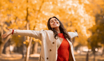 Cheerful young woman in fashionable fall outfit feeling free and enjoying herself at autumn park, panorama