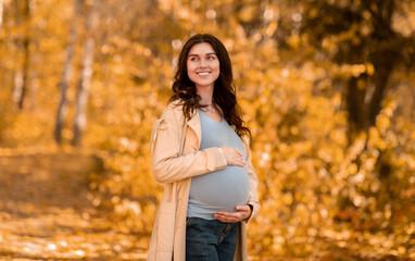 Beautiful young pregnant woman hugging her belly on walk in autumn park