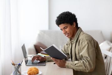 E-learning concept. Handsome black teenager using his notes to make online home assignment on laptop indoors