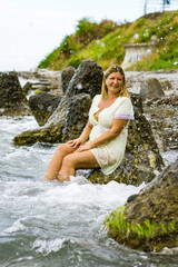A young blonde woman in a short yellow dress sits on large stones, knee-deep in water on the seashore.