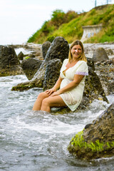 A young blonde woman in a short yellow dress sits on large stones, knee-deep in water on the seashore.