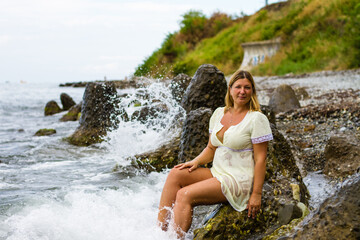 A young blonde woman in a short yellow dress sits on large stones, knee-deep in water on the seashore.