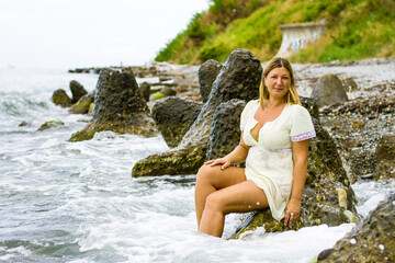 A young blonde woman in a short yellow dress sits on large stones, knee-deep in water on the seashore.
